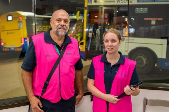 Male and female CLO wearing bright pink uniform vests, standing at the bus station with buses behind